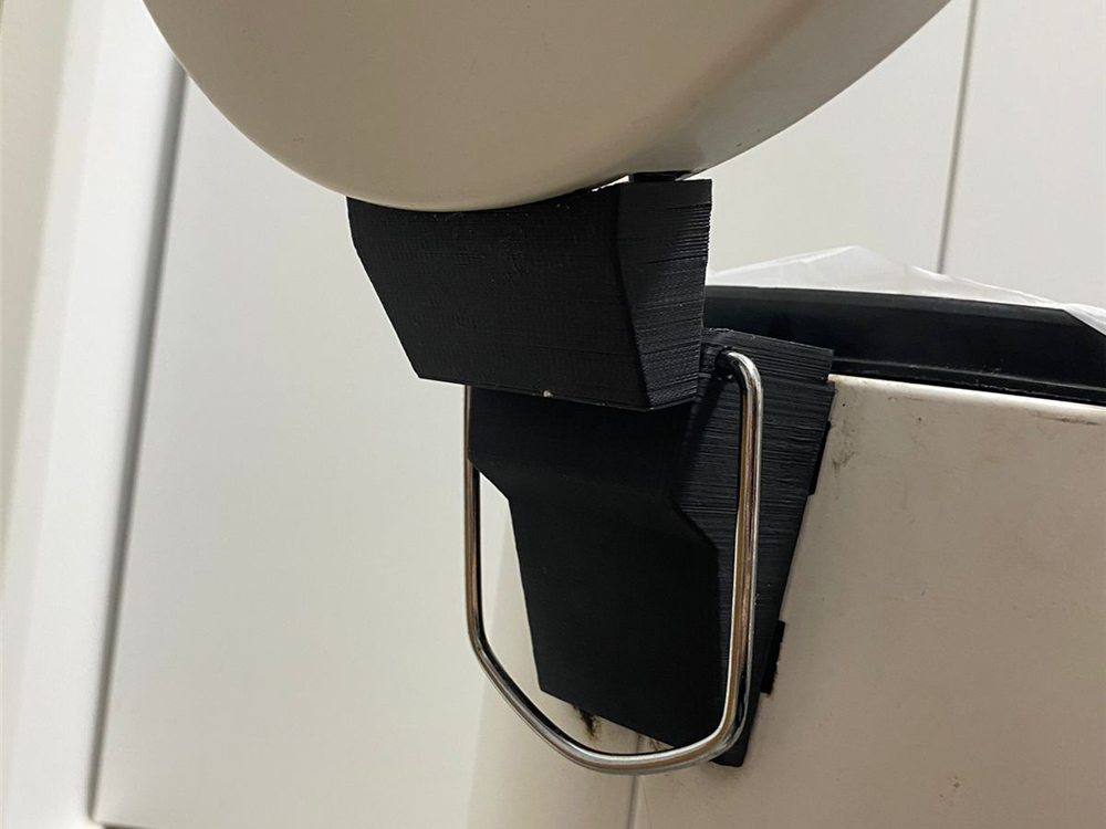 Mecanismo negro impreso en 3D montado en la puerta de un mueble de cocina, enganchado al asa de alambre de un tacho de basura.