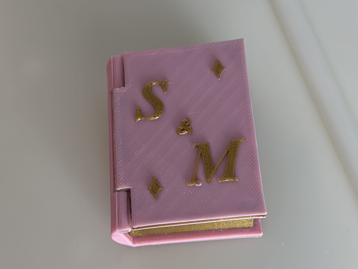 Caja de anillo con forma de libro, impresa en 3D en rosa y dorado, con dos iniciales en relieve en la tapa.