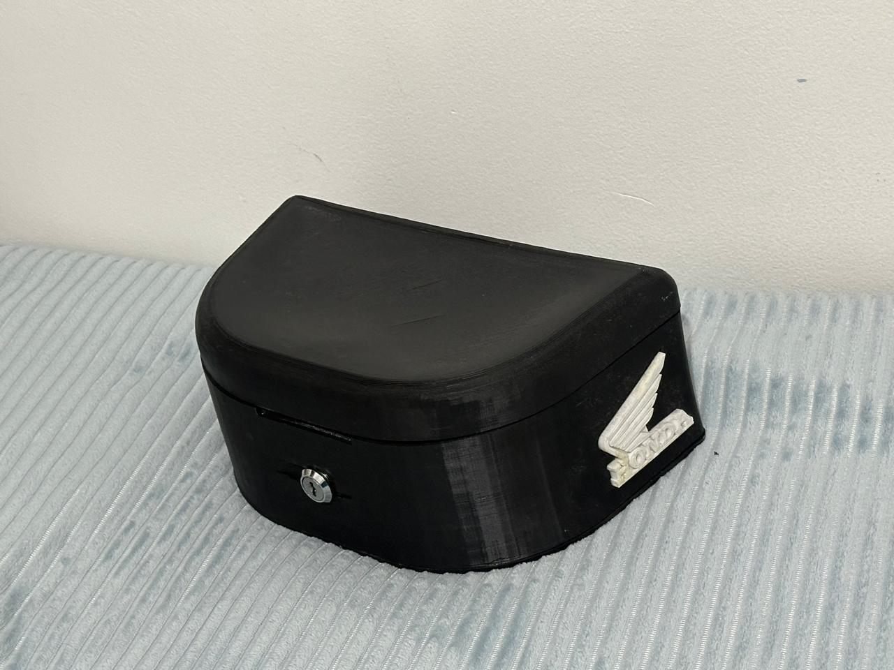 El baúl para la Honda GLH 150 impreso en 3D en negro, cerrado, con la cerradura al frente y el ala de Honda en blanco.